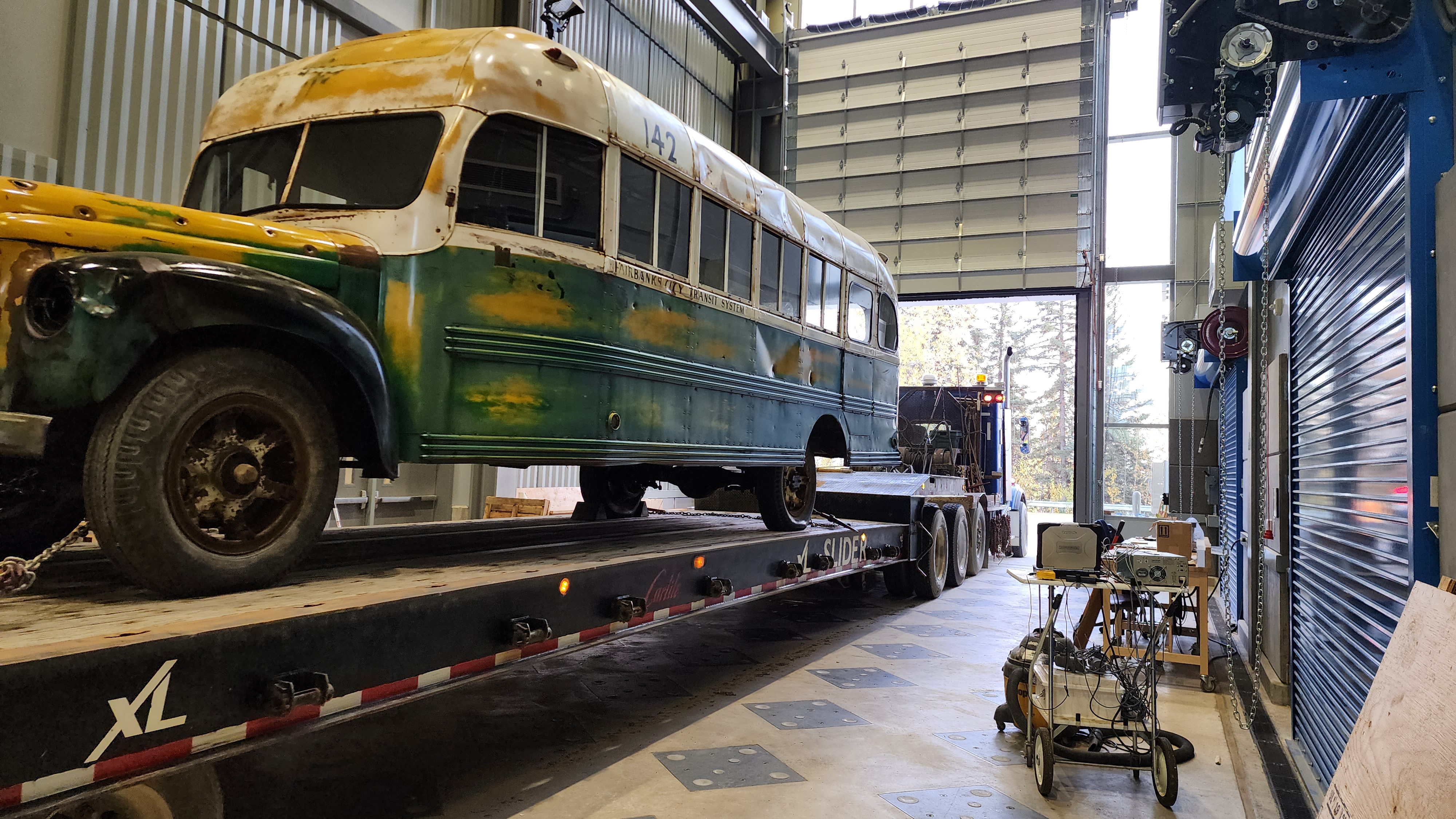 Bus 142 sits on a flatbed trailer inside a large workshop bay.
